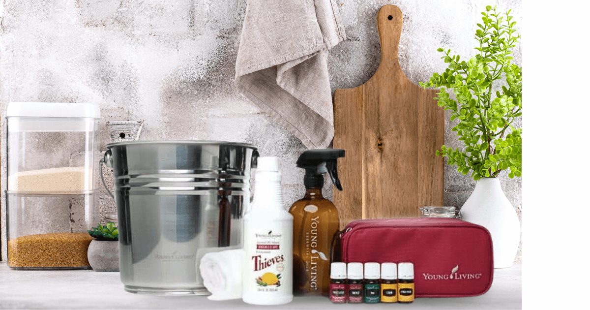 A white marble kitchen counter with misc cutting board, spice container, towel and plant behind and a stainless steel bucket, towel, thieves household cleaner, spray bottle, red essential oil storage pouch and 5 young living essential oils next to it for a Bound to Journal Blog post titled Thieves Household Cleaner: Your Year-Round Solution for Safe, Natural Cleaning