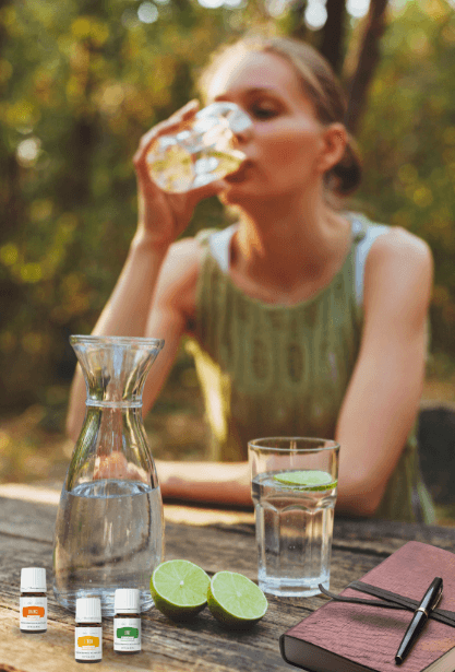  Woman sitting at a picnic table drinking water next to a caraf of water, cut limes, a water glass with a slice of lime, a journal & pen, and 3 bottles of Young Living vitality essential oils  for a Bound to Journal blog post titled Add Vitality: Essential Oils to Make Hydration and Summer Journaling More Fun