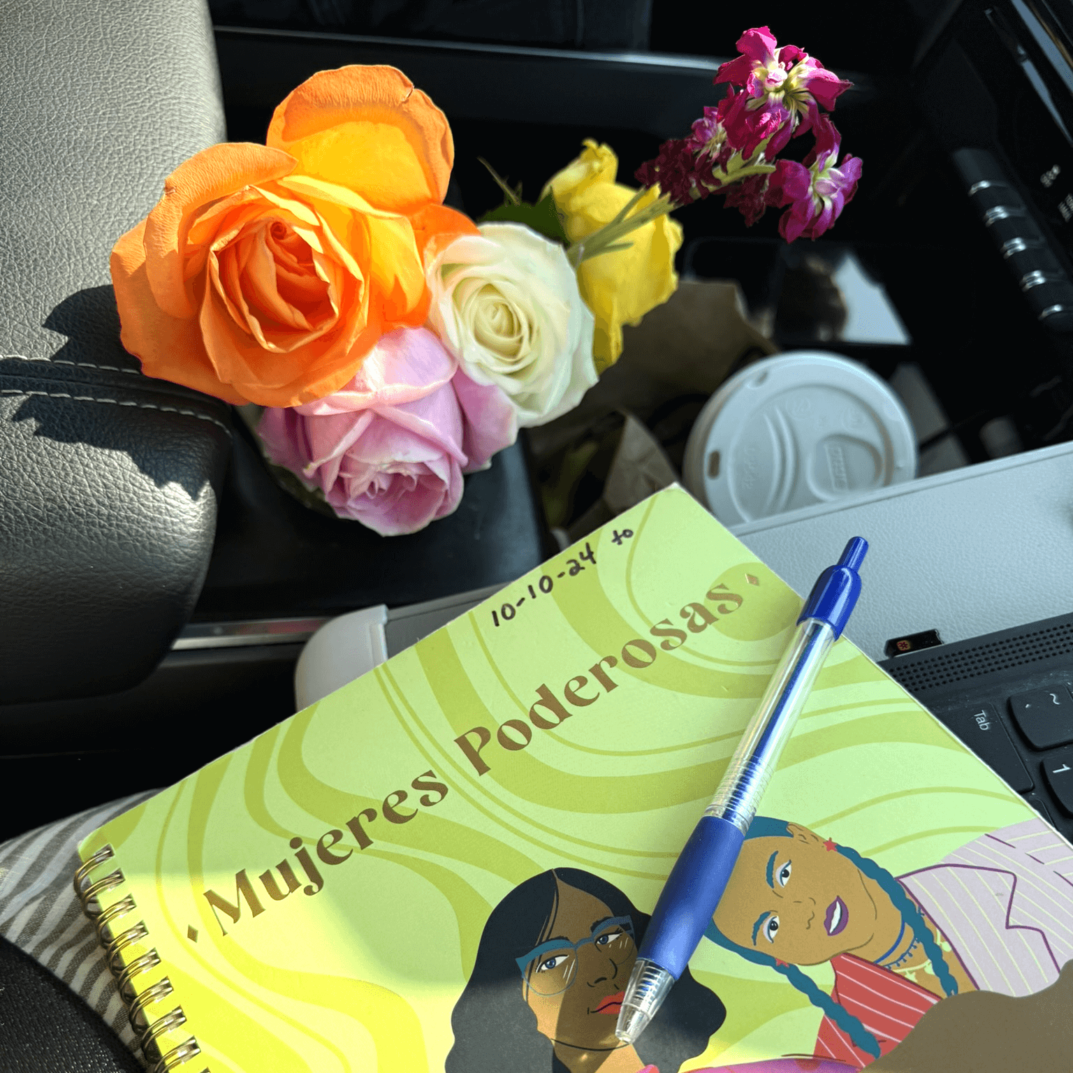 Fresh roses in the council of the vehicle next to the lid of a to-go coffee cup and a Mujeres Ponderosas daily journal and pen for a Bound to Journal blog post titled Amplifying the Benefits of Journaling: Engaging Your Senses