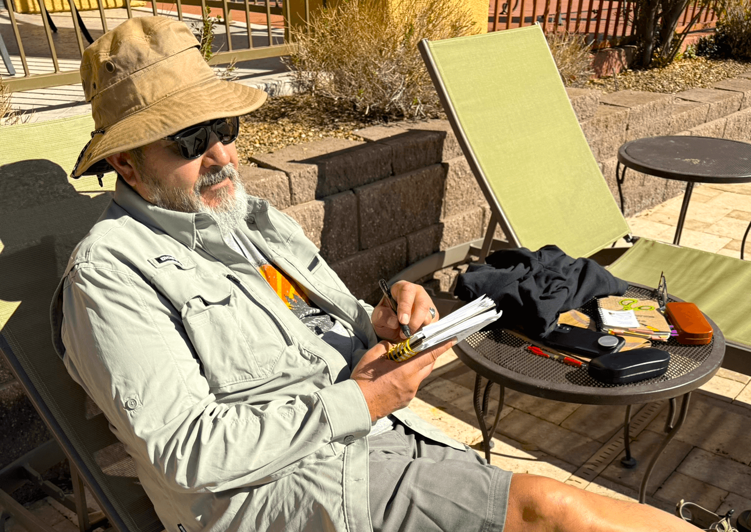 Allen from Bound to Journal wearing sunglasses and hat journaling while sitting on a lounge chair next to a table with a journal and sunglasses appears poolside for a Bound to Journal blog post titled Amplifying the Benefits of Journaling: Engaging Your Senses