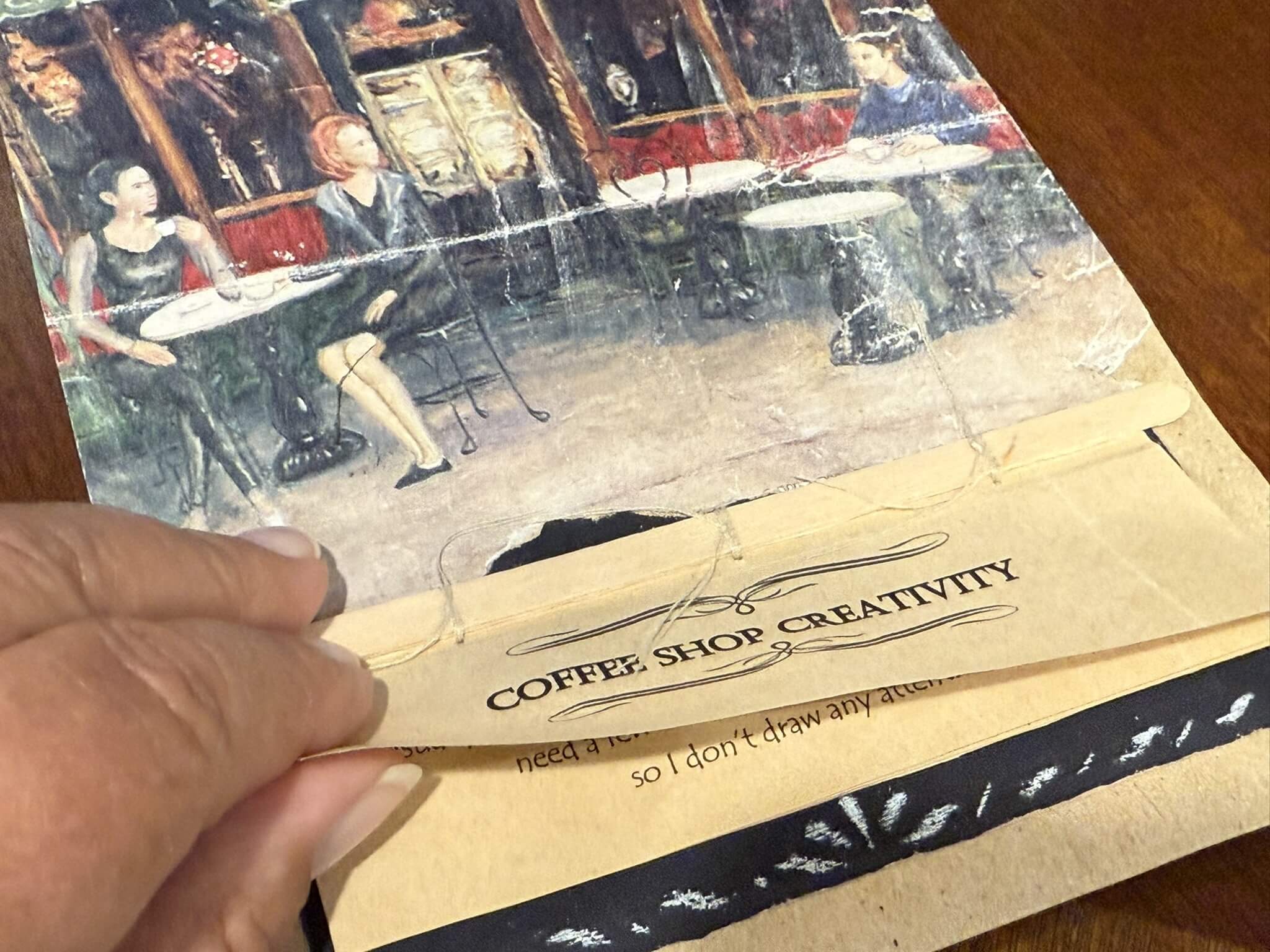 Fingers turning the attatched page of a coffee shop with the image of two ladies sitting at an outdoor cafe for a Bound to Journal blog post titled Amplifying the Benefits of Journaling: Engaging Your SensesLetter to Mary