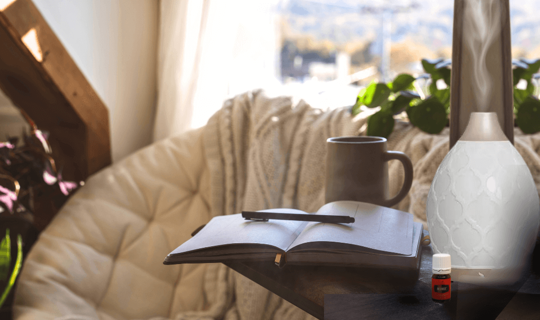  An open journal, pen, bottle of Young Living essential oil, a diffuser, and a coffee cup on an end table next to a chair, by plants, and a window overlooking nature  for a new Bound to Journal blog post titled Creating a Healthy Home: Improving Air Quality for a Safer, Rejuvenating Space