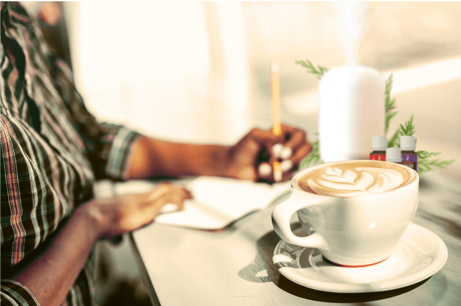  A person sitting at the table journaling next to a cup of coffee with a beautiful froth design next to a diffuser and essential oils