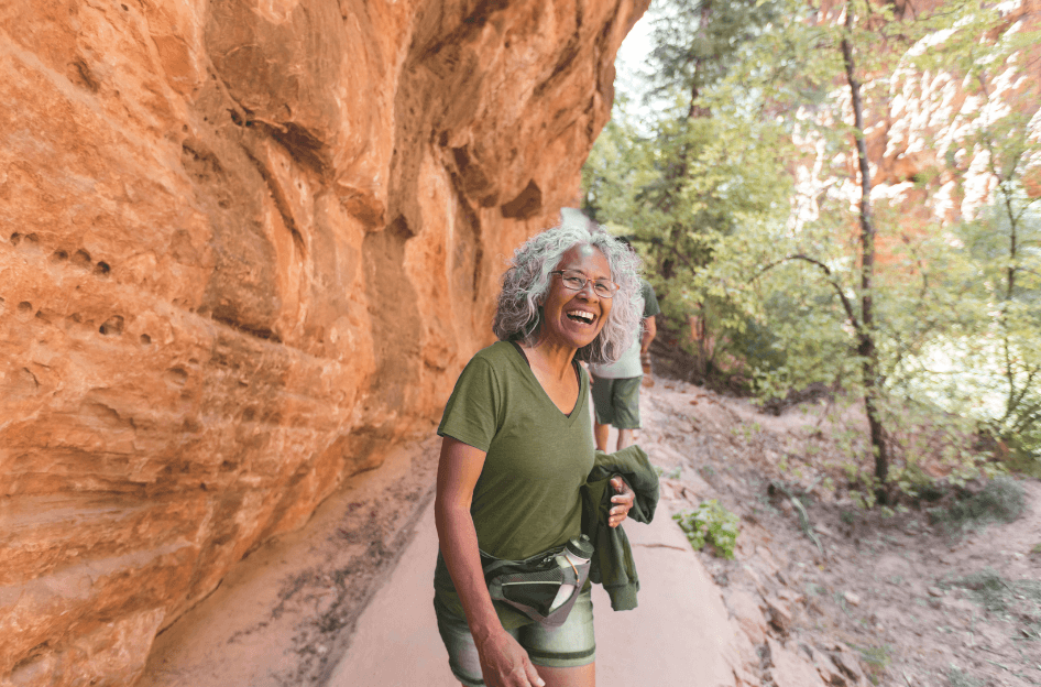  A woman pausing to behind her hiking partners, wearing hiking clothes and a big smile on a nature hike next to a large rock face  for a Bound to Journal blog post titled Reignite Your Passions: Journaling & Creativity for Aging Gracefully