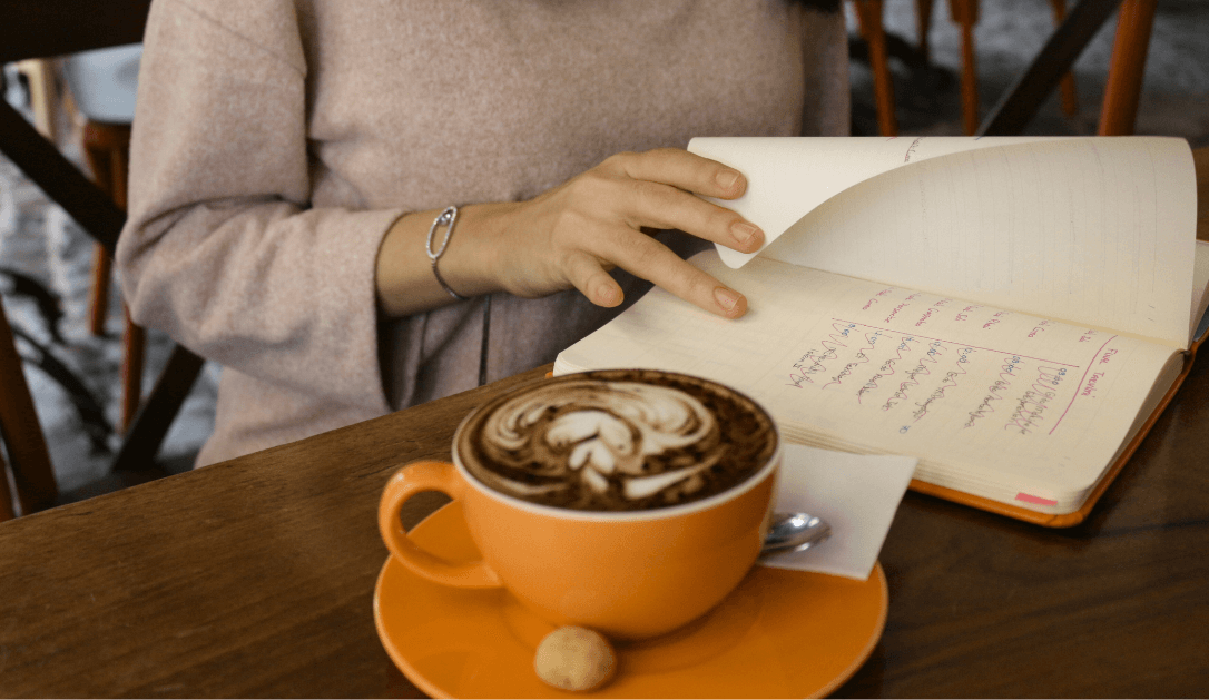  A woman sitting at a table with an open journal and a bright orange cup and saucer with a steamed milk design  for a Bound to Journal blog post titled Reignite Your Passions: Journaling & Creativity for Aging Gracefully