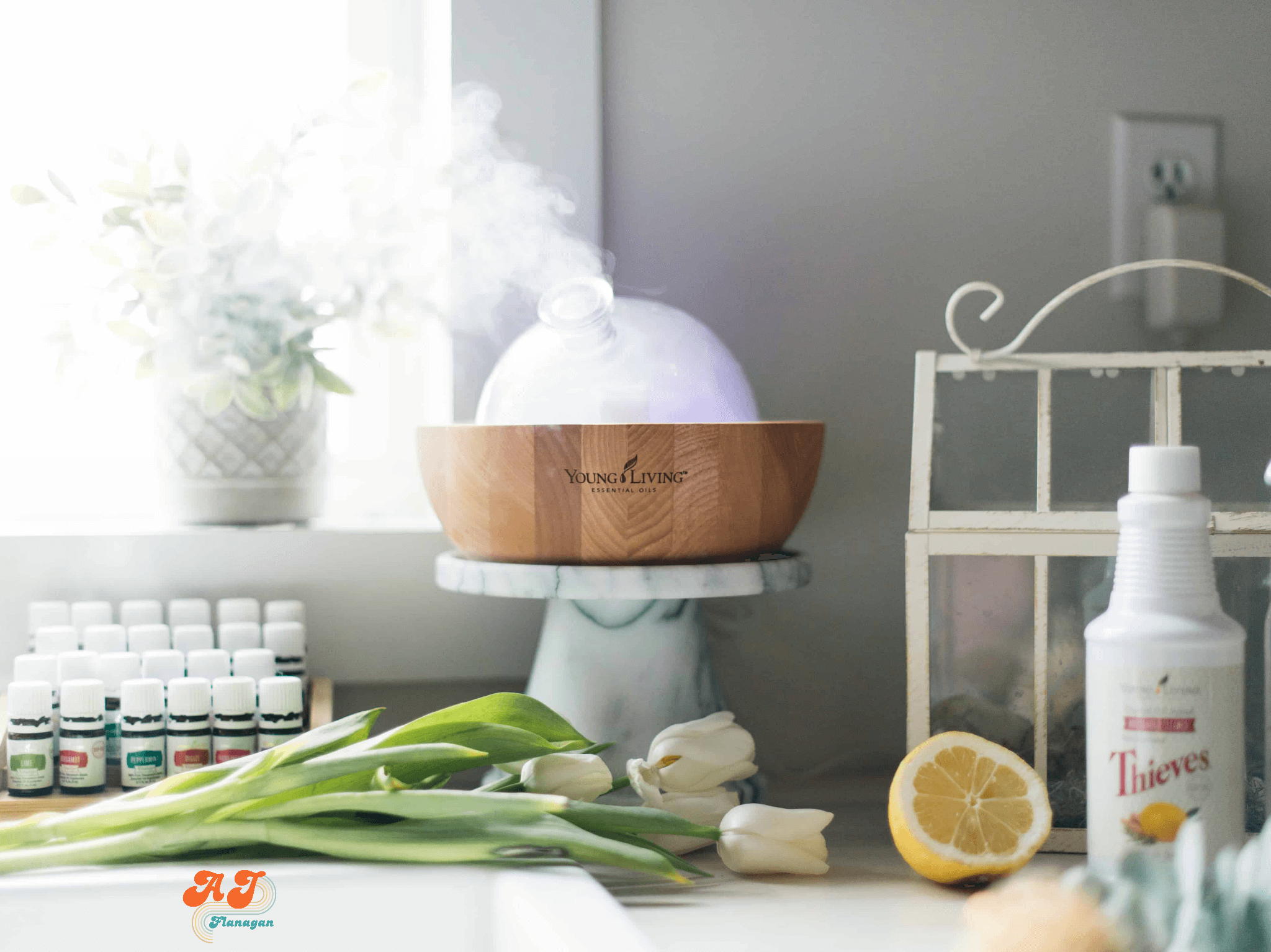 A bright and airy photo of a white kitchen counter with essential oils, a diffuser, a fresh lemon, white tulips, and a bottle of Thieves Household Cleaner on it