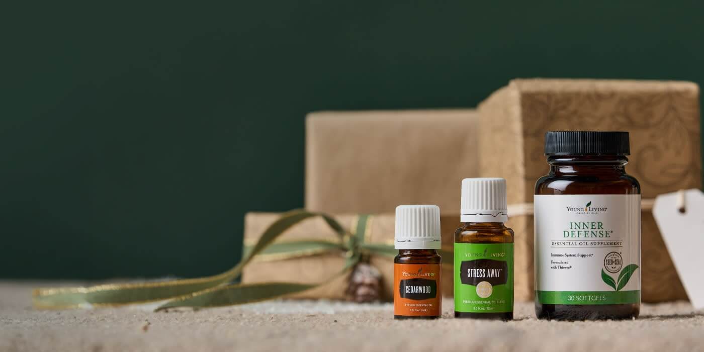 Young Living® Cedarwood™, Stress Away™, and Inner Defense® arranged in a warm holiday scene with wrapped gifts, soft green ribbon, and a neutral background—simple, festive product imagery for December.