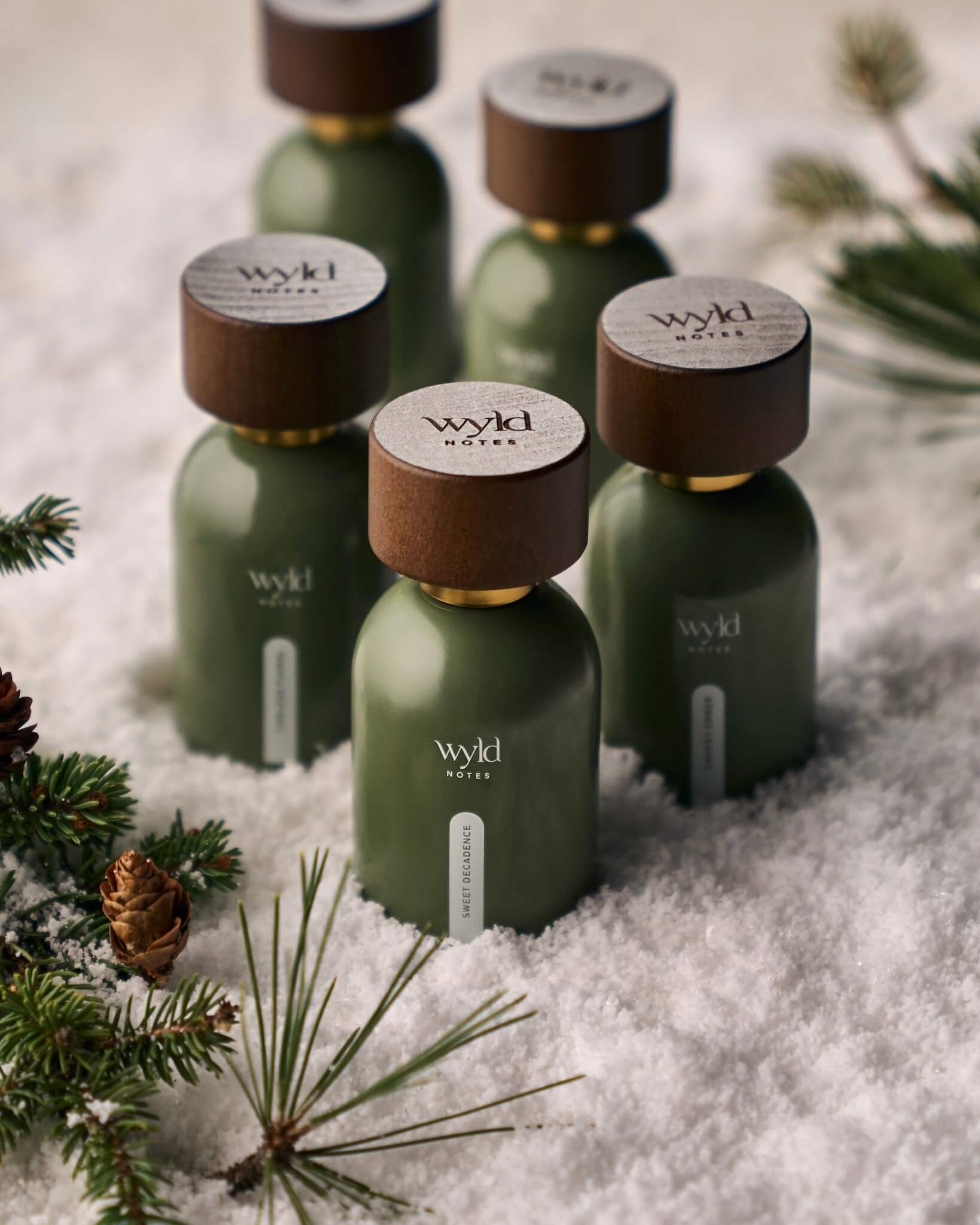 Five Wyld Notes® botanical fine fragrances arranged in a snowy winter scene with frosted wood caps. A clean, gender-neutral collection inspired by forest, citrus, floral, earthy, and warm natural elements — perfect for seasonal gifting.