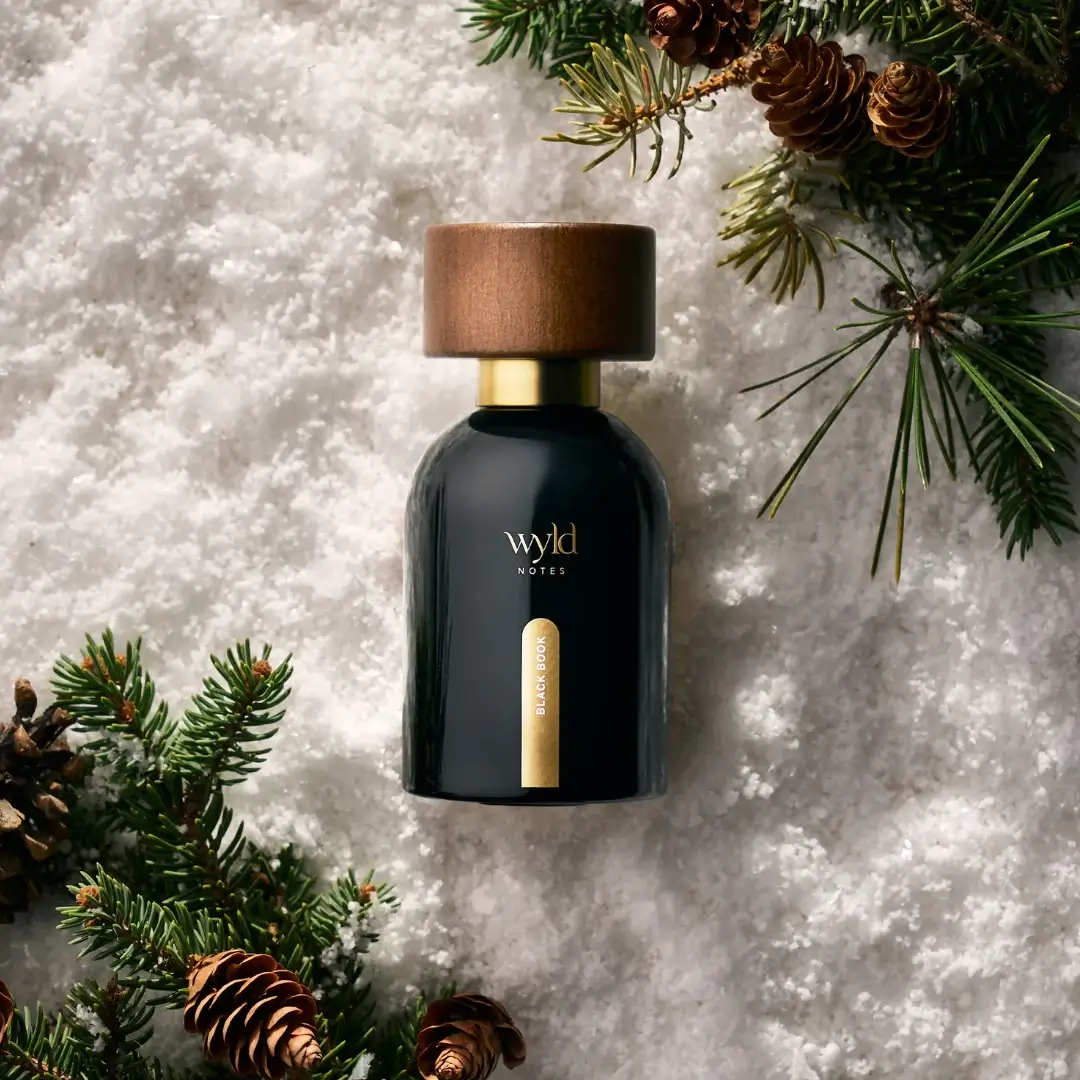 Wyld Notes™ Black Book™ clean botanical fragrance photographed in a winter scene with snow, pine branches, and warm shadows — nontoxic gender-neutral vegan perfume with deep woody notes, luxe holiday gift idea.