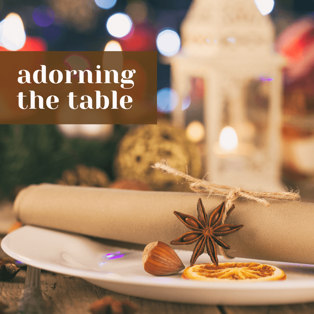  Holiday place setting with a rolled napkin tied in twine, star anise, hazelnut, and dried orange slice on a white plate, with glowing candles in the background and the words ‘adorning the table.